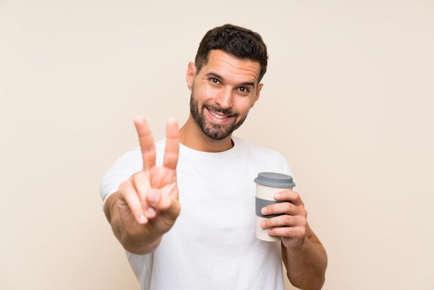 Giovane con la barba che tiene un caffè da asporto sopra la parete blu isolata che sorride e che mostra il segno di vittoria