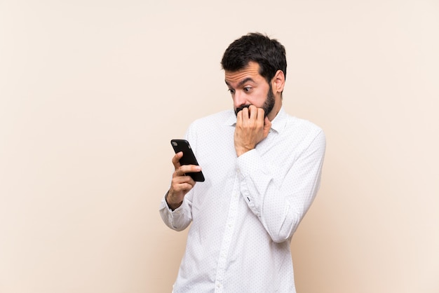 Giovane con la barba che tiene un cellulare nervoso e spaventato mettendo le mani alla bocca