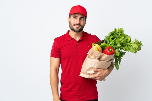 Giovane con la barba che tiene un sacchetto pieno di verdure isolato su sfondo bianco in piedi e guardando al lato