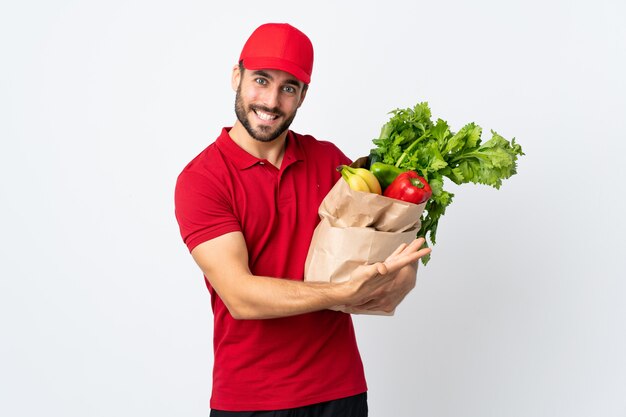 Giovane con la barba che tiene una borsa piena di verdure isolate sulla parete bianca che estende le mani al lato per invitare a venire