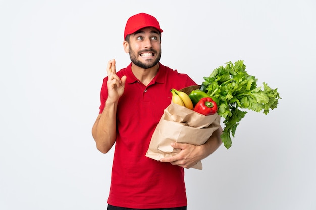 Giovane con la barba che tiene una borsa piena di verdure isolate sulla parete bianca con l'incrocio delle dita