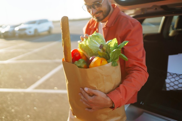 Giovane con la borsa della spesa piena di verdure vicino all'auto Bell'uomo dopo lo shopping