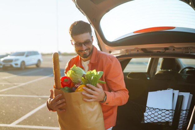 Giovane con la borsa della spesa piena di verdure vicino all'auto Bell'uomo dopo lo shopping