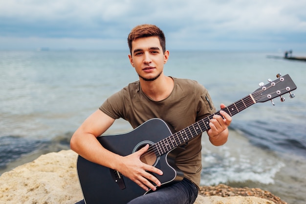 Giovane con la chitarra acustica che gioca sulla spiaggia circondata con le rocce il giorno piovoso