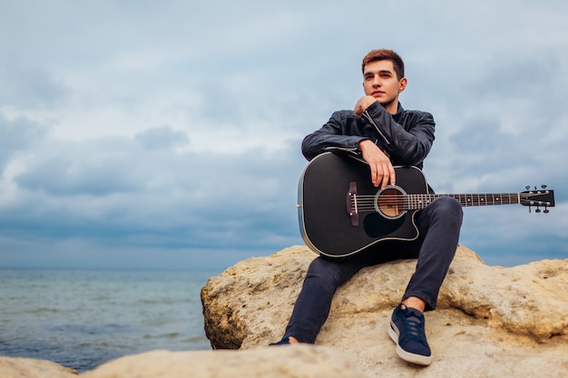 Giovane con la chitarra acustica che si siede sulla spiaggia circondata con le rocce il giorno piovoso