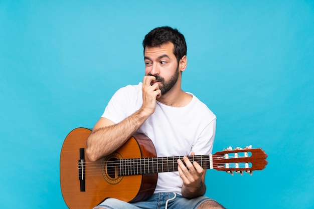 Giovane con la chitarra sopra la parete blu isolata che ha dubbi