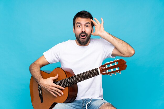 Giovane con la chitarra sopra la parete blu isolata con l'espressione di sorpresa