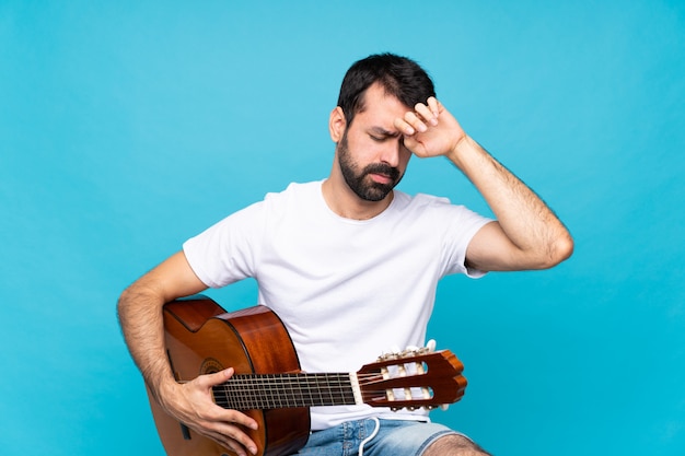 Giovane con la chitarra sopra la parete blu isolata con l'espressione stanca e malata