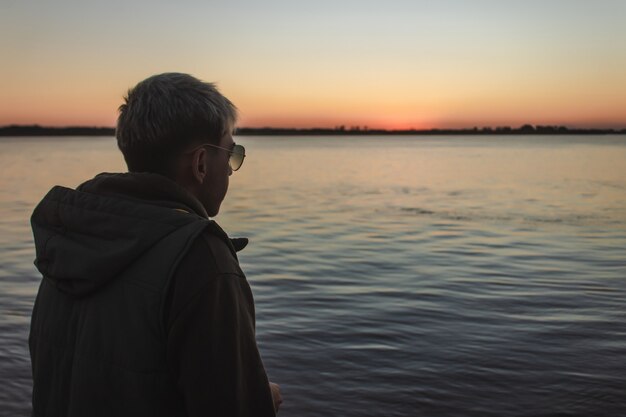 Giovane con occhiali da sole a guardare il tramonto in riva al mare.