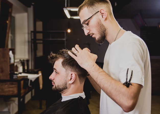 Giovane con taglio di capelli alla moda al negozio di barbiere. Il barbiere fa la pettinatura e la barba.