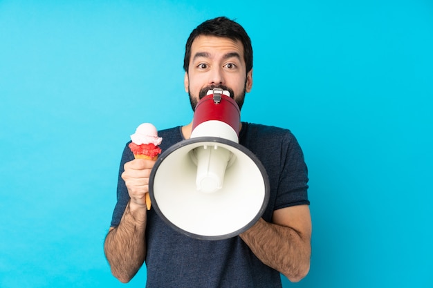 Giovane con un gelato della cornetta sopra la parete blu isolata che grida tramite un megafono