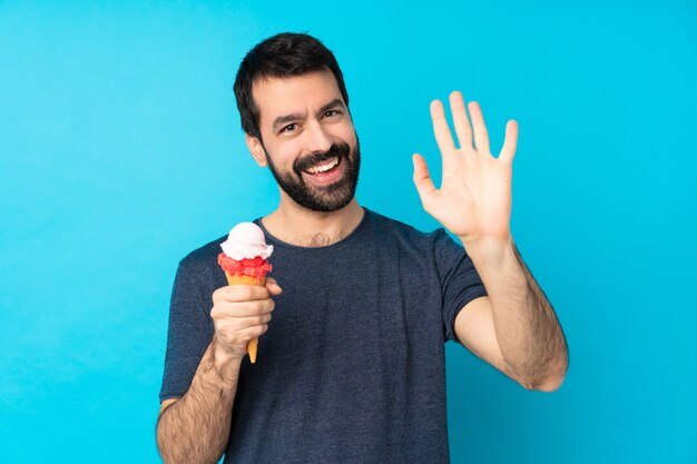 Giovane con un gelato della cornetta sopra la parete blu isolata che saluta con la mano con l'espressione felice