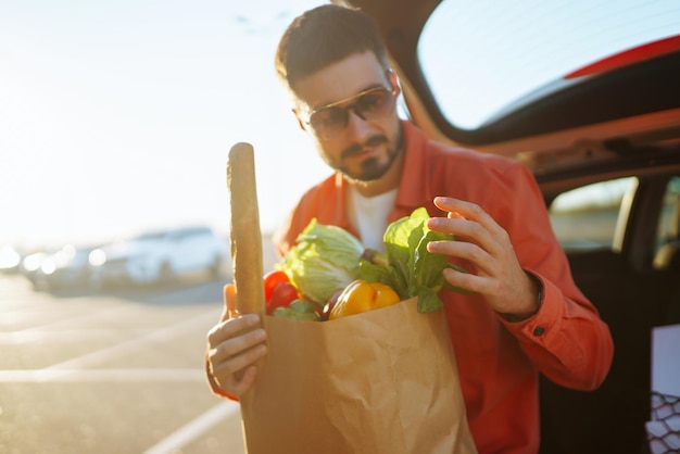 Giovane con una borsa piena di verdure vicino all'auto Uomo bello dopo aver fatto la spesa