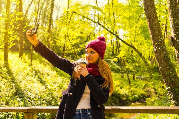 Giovane con una giacca nera, una sciarpa e un cappello di lana rosso che si diverte in un parco autunnale, facendo un selfie con il cellulare