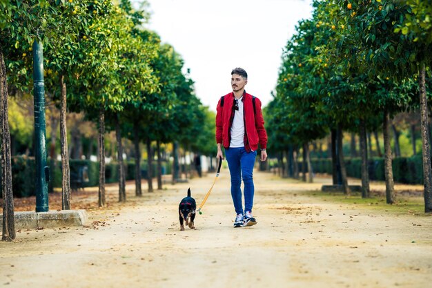 Giovane con una giacca rossa che porta a spasso un cane di piccola taglia in un parco urbano