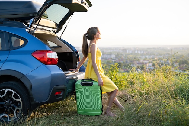 Giovane conducente femminile che riposa seduto su una valigia vicino alla sua auto in campo estivo Concetto di viaggio e vacanze