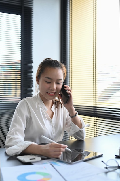 Giovane consulente finanziario femminile che ha una conversazione telefonica con il cliente e utilizza la tavoletta digitale sul posto di lavoro