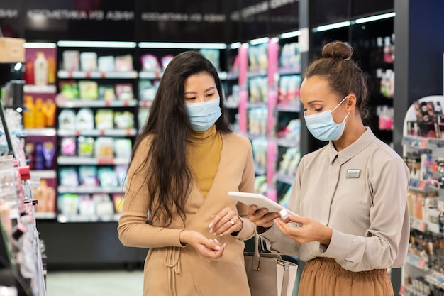 Giovane consulente in maschera protettiva che scorre nel tablet mentre cerca il prezzo del lucidalabbra ricercato da uno dei clienti nel supermercato di bellezza