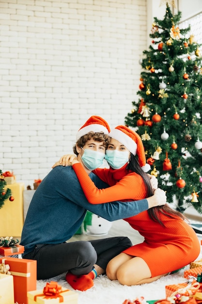 Giovane coppia allegra in cappelli di Babbo Natale e indossando maschere mediche in quarantena seduti circondati da scatole regalo colorate che si divertono a celebrare il Natale a casa.