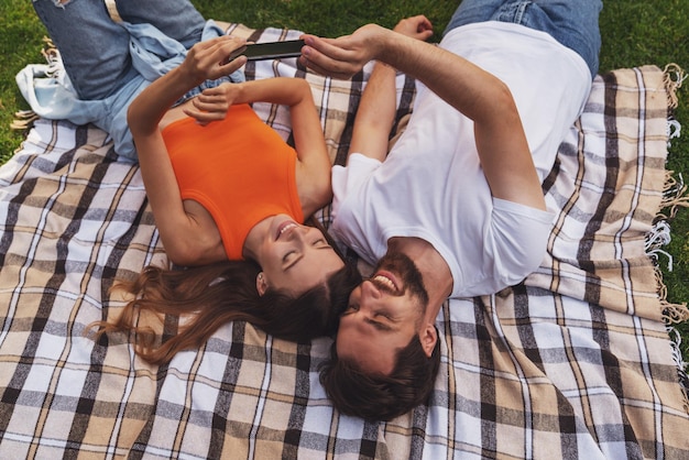 Giovane coppia allegra sdraiata su una coperta nel parco durante un picnic e facendo un selfie usando il telefono.