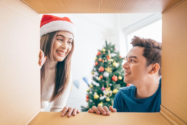 Giovane coppia asiatica che apre un regalo di Natale insieme con un sorriso viste dall'interno della scatola con un albero di Natale decorato sullo sfondo Santo Stefano durante le vacanze di Natale