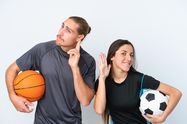 Giovane coppia caucasica che gioca a basket e calcio isolati su sfondo bianco ascoltando qualcosa mettendo la mano sull'orecchio