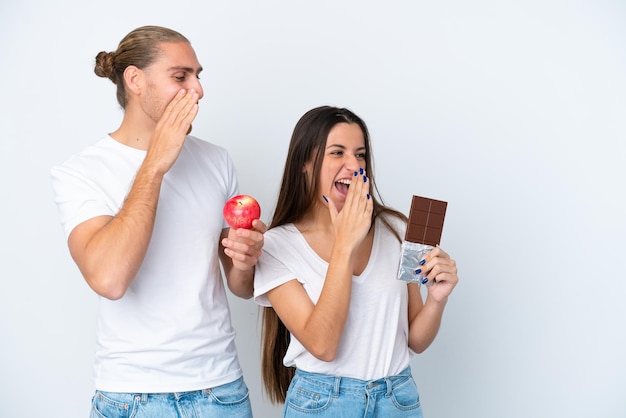 Giovane coppia caucasica con cioccolato e mela isolata su sfondo bianco che grida con la bocca spalancata lateralmente