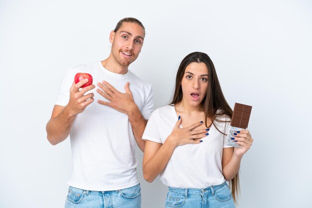 Giovane coppia caucasica con cioccolato e mela isolata su sfondo bianco sorpresa e scioccata mentre guarda a destra