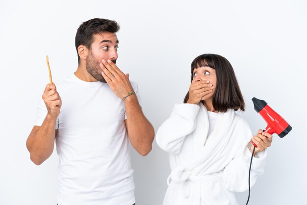Giovane coppia caucasica in possesso di un asciugacapelli e spazzolino da denti isolati su sfondo bianco che copre la bocca con le mani per dire qualcosa di inappropriato