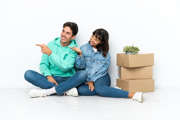 Giovane coppia che fa una mossa mentre prende una scatola piena di cose seduto sul pavimento isolato su sfondo bianco che punta indietro e presenta un prodotto