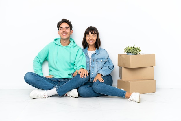 Giovane coppia che fa una mossa mentre prende una scatola piena di cose seduto sul pavimento isolato su sfondo bianco in posa con le braccia all'anca e sorridente