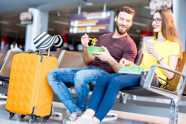 Giovane coppia che fa uno spuntino con cestini per il pranzo nella sala d'attesa dell'aeroporto durante la vacanza