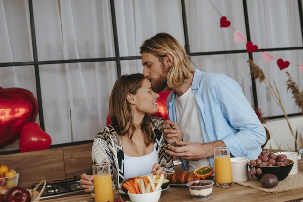 Giovane coppia che gusta la colazione in cucina insieme a palloncini a forma di cuore rosso sullo sfondo