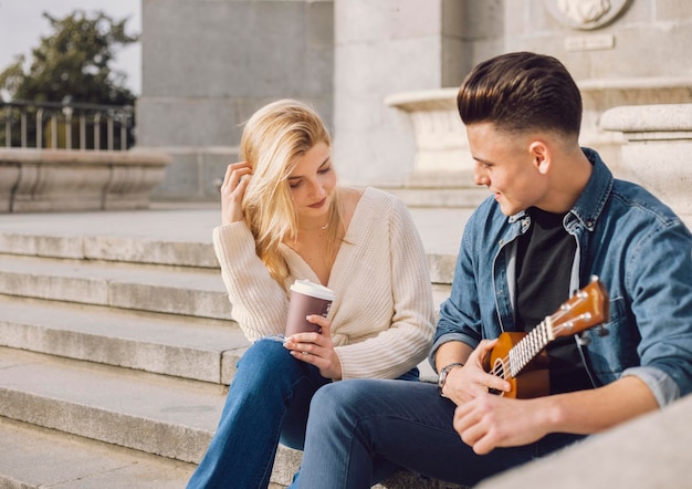 Giovane coppia che ha un appuntamento in città Sono seduti Lui ha un ukulele e lei tiene un caffè