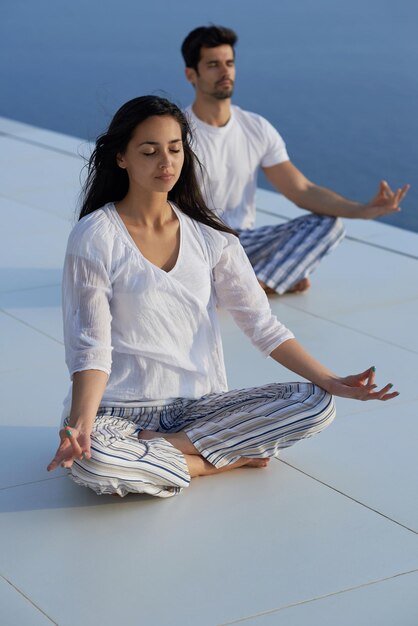 giovane coppia che pratica yoga al tramonto nella moderna terrazza domestica con l'oceano e il tramonto sullo sfondo