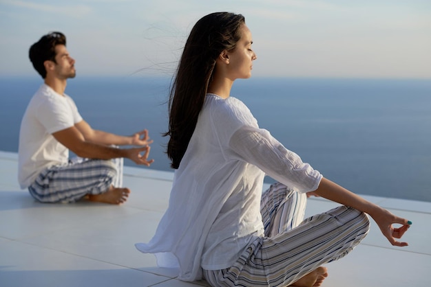 giovane coppia che pratica yoga al tramonto nella moderna terrazza domestica con l'oceano e il tramonto sullo sfondo