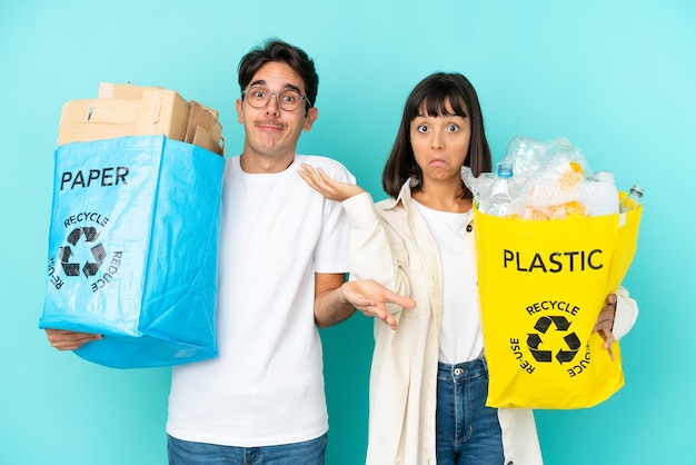 Giovane coppia che tiene un sacchetto pieno di plastica e carta da riciclare isolato su sfondo blu che ha dubbi mentre alza le mani e le spalle