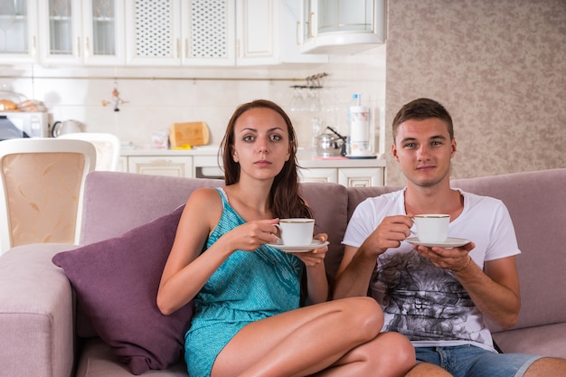 Giovane coppia con tazze di caffè o tè seduti insieme sul divano e fissando la telecamera - godersi il caffè mattutino insieme a casa