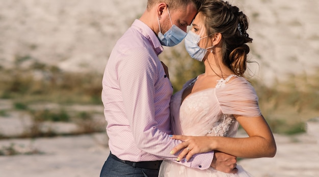 Giovane coppia di innamorati che cammina con maschere mediche nel parco durante la quarantena il giorno del loro matrimonio.