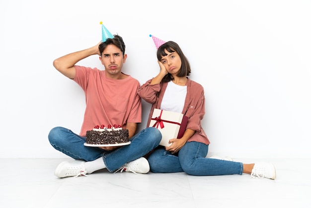 Giovane coppia di razza mista che celebra un compleanno seduto sul pavimento isolato su sfondo bianco con un'espressione di frustrazione e di non comprensione