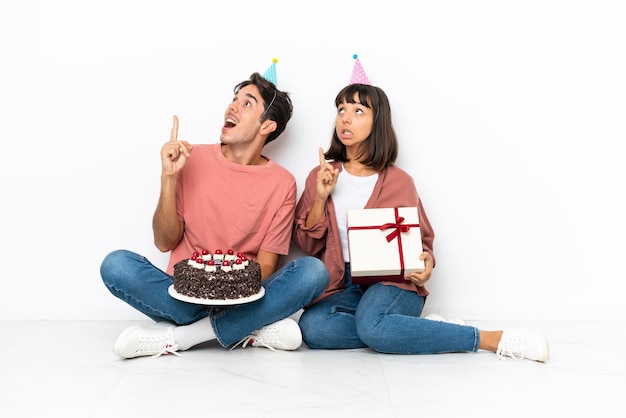 Giovane coppia di razza mista che celebra un compleanno seduto sul pavimento isolato su sfondo bianco pensando a un'idea che punta il dito verso l'alto