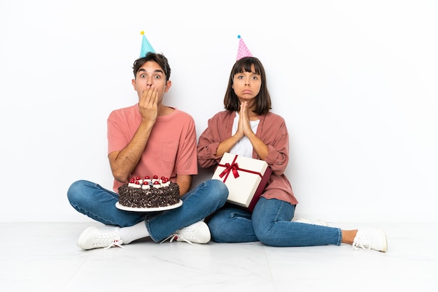 Giovane coppia di razza mista che celebra un compleanno seduto sul pavimento isolato su sfondo bianco tiene insieme il palmo La persona chiede qualcosa