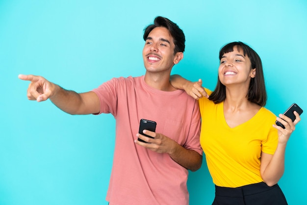 Giovane coppia di razza mista che tiene il telefono cellulare isolato su sfondo blu che presenta un'idea mentre guarda sorridendo verso