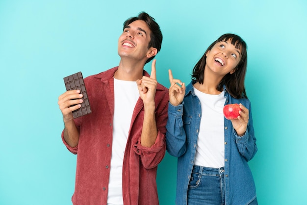 Giovane coppia di razza mista che tiene mela e cioccolato isolati su sfondo blu che punta con il dito indice una grande idea