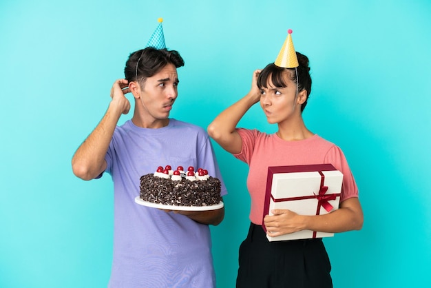 Giovane coppia di razza mista che tiene torta di compleanno e presente isolato su sfondo blu che ha dubbi mentre si gratta la testa