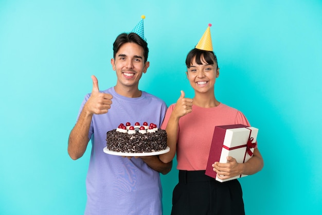 Giovane coppia di razza mista che tiene torta di compleanno e presente isolato su sfondo blu dando un pollice in alto gesto con entrambe le mani e sorridente