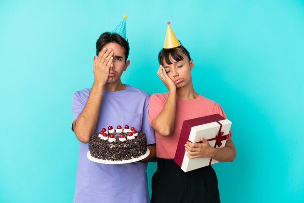 Giovane coppia di razza mista che tiene torta di compleanno e regalo isolato su sfondo blu con espressione facciale sorpresa e scioccata