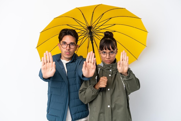 Giovane coppia di razza mista che tiene un ombrello isolato su sfondo bianco facendo un gesto di arresto per deluso da un'opinione