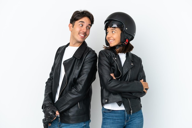 Giovane coppia di razza mista con un casco da motociclista isolato su sfondo bianco guardando sopra la spalla con un sorriso
