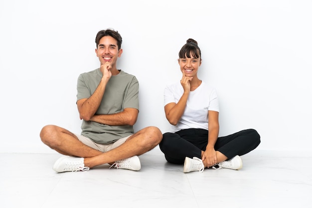 Giovane coppia di razza mista seduta sul pavimento isolato su sfondo bianco sorridente con un'espressione dolce